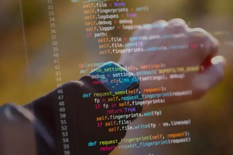 A hand wearing a watch, with code on a monitor in the background.